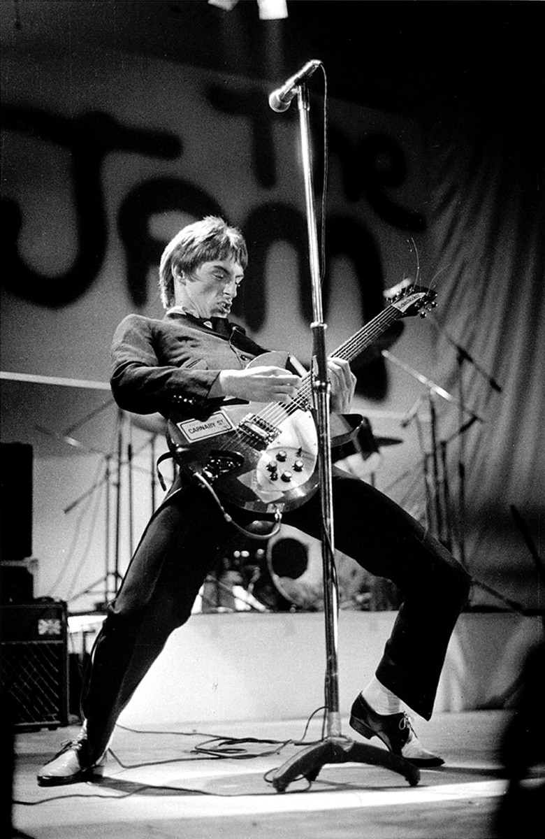 Paul Weller performing live onstage, playing Rickenbacker 330 guitar, with The Jam logo on backdrop behind, 